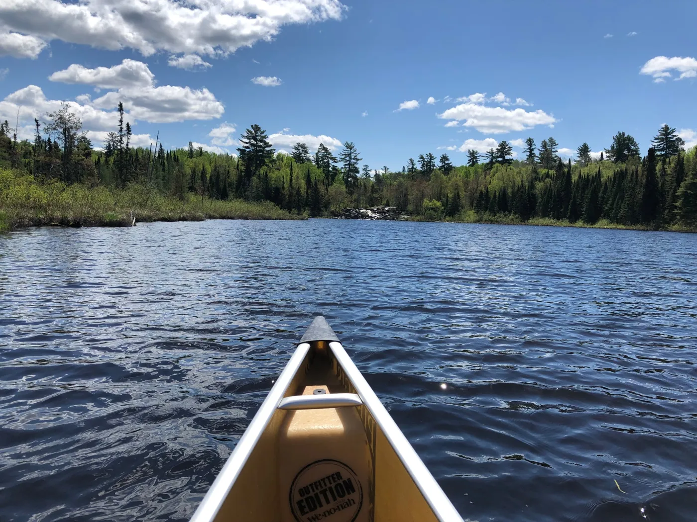 View from the bow of a canoe across a Boundary Waters lake toward a small waterfall and a pine-lined shore