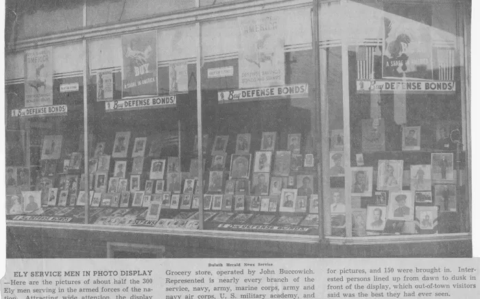 A store window filled with framed photos of Ely servicemen under Buy Defense Bonds signs, with a Duluth Herald caption below