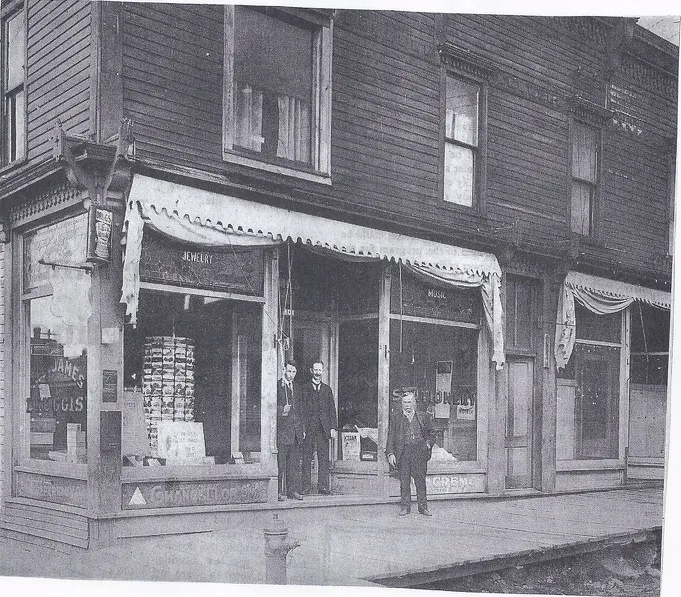 The wood-frame storefront with James Druggist, jewelry, music and stationery signs and three men out front