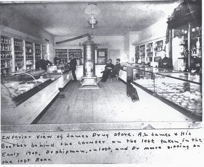 Interior of James Drug Store in the early 1900s, with glass display cases, shelves of bottles and a tall stove in the middle of the room