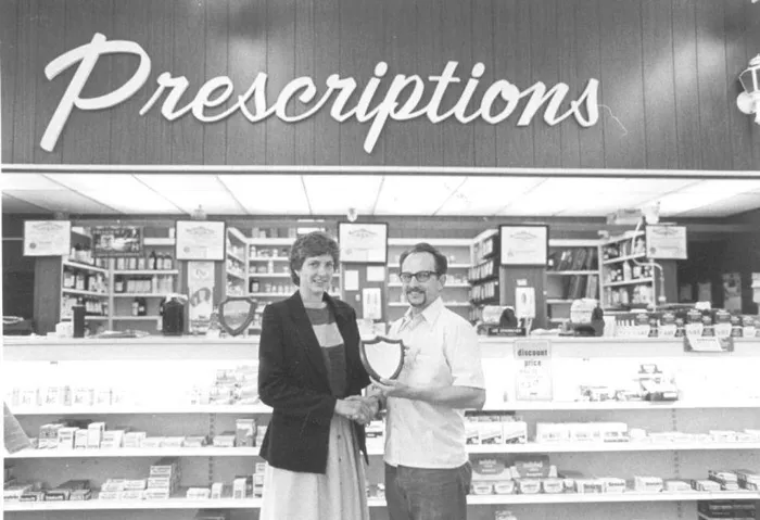 A woman and a man shaking hands and holding a plaque in front of the Prescriptions counter