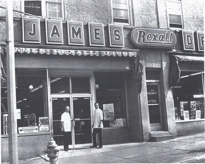 Two pharmacists in white coats standing at the glass doors of James Rexall Drugs at number 101