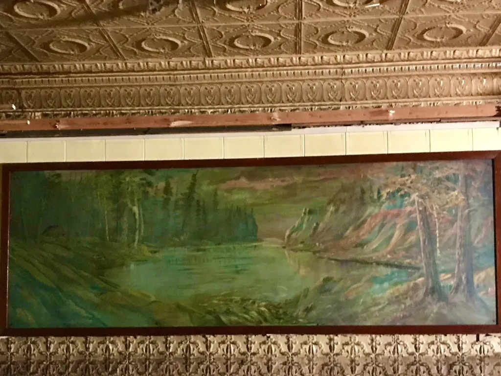 An old framed landscape painting of a North Woods lake beneath tin cornice and wall panels