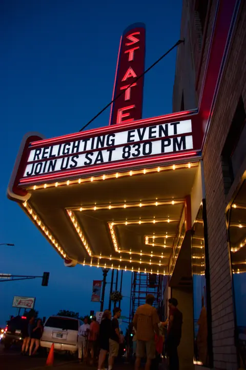 The restored marquee lit at night reading Relighting Event, Join Us Sat 830 PM