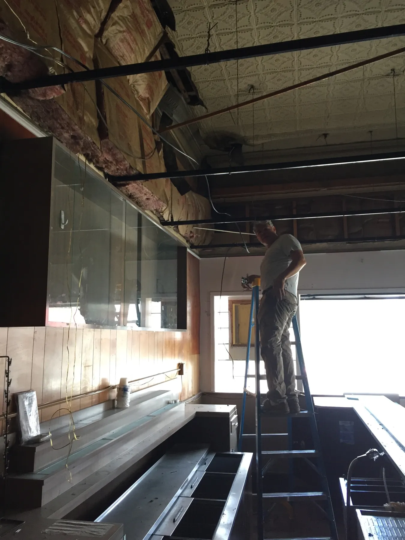 A man on a ladder beside the old bar with the drop ceiling removed and tin ceiling showing above